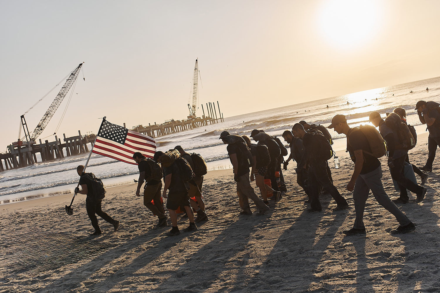 Group of people with backpacks walk on a beach at sunset, one carrying an American flag; cranes in background.