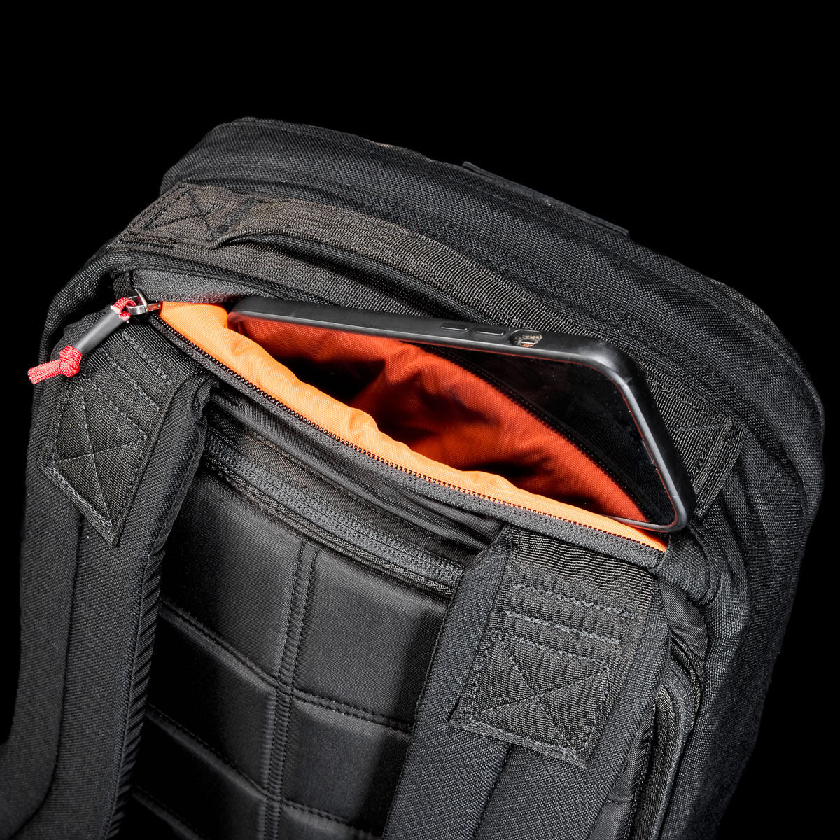 The GR1 Shooter 26L - Built by Scars backpack is made from rugged CORDURA®, features an orange lining, and has a zippered pocket near the shoulder straps for convenient phone storage.