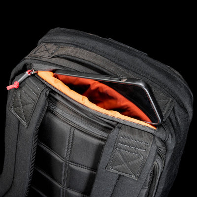 The GR1 Shooter 26L - Built by Scars backpack is made from rugged CORDURA®, features an orange lining, and has a zippered pocket near the shoulder straps for convenient phone storage.