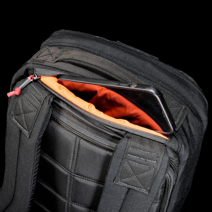 The GR1 Shooter 26L - Built by Scars backpack is made from rugged CORDURA®, features an orange lining, and has a zippered pocket near the shoulder straps for convenient phone storage.