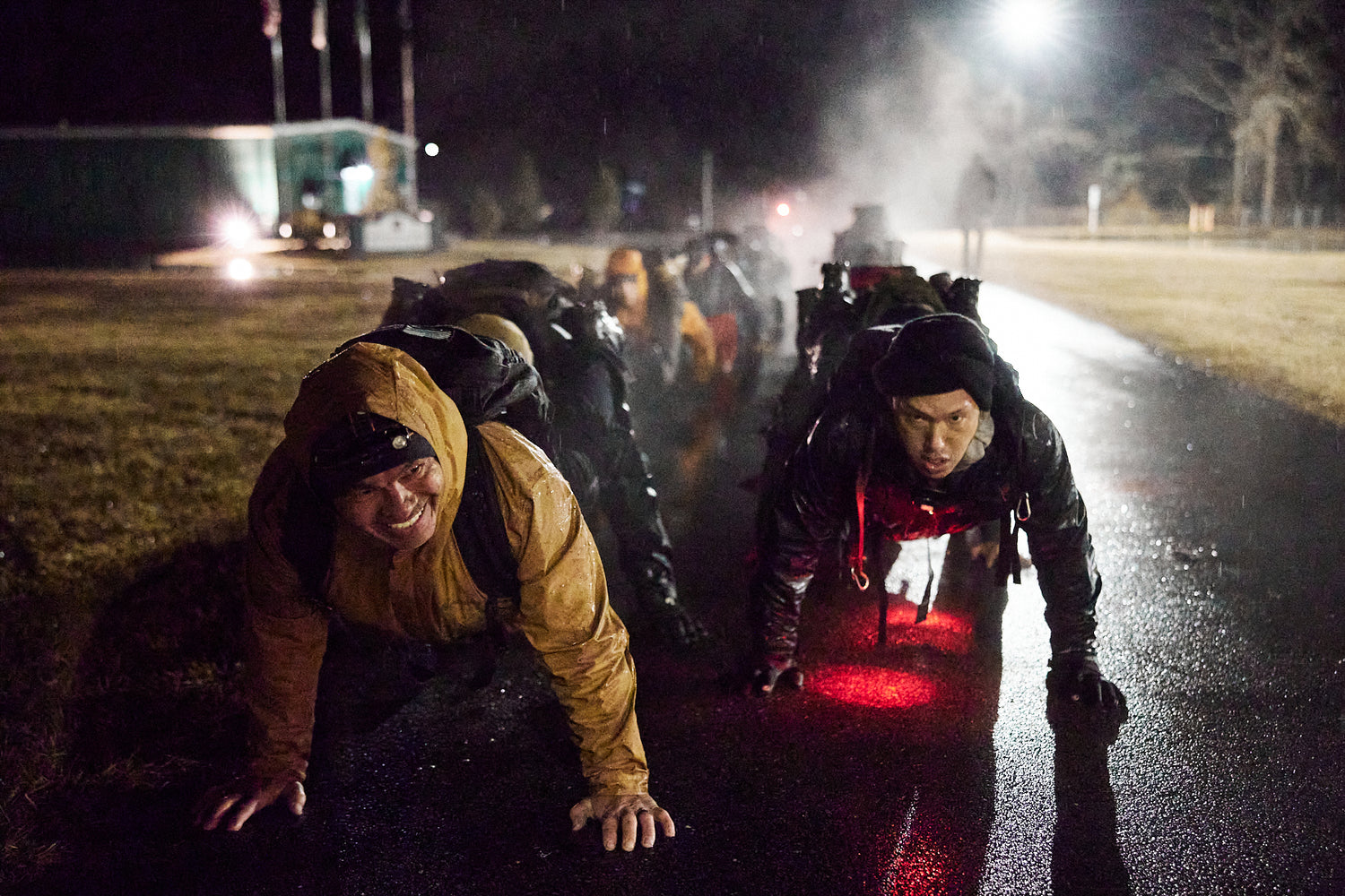 People in outdoor gear crawl on wet pavement at night, illuminated by red lights and street lamps.
