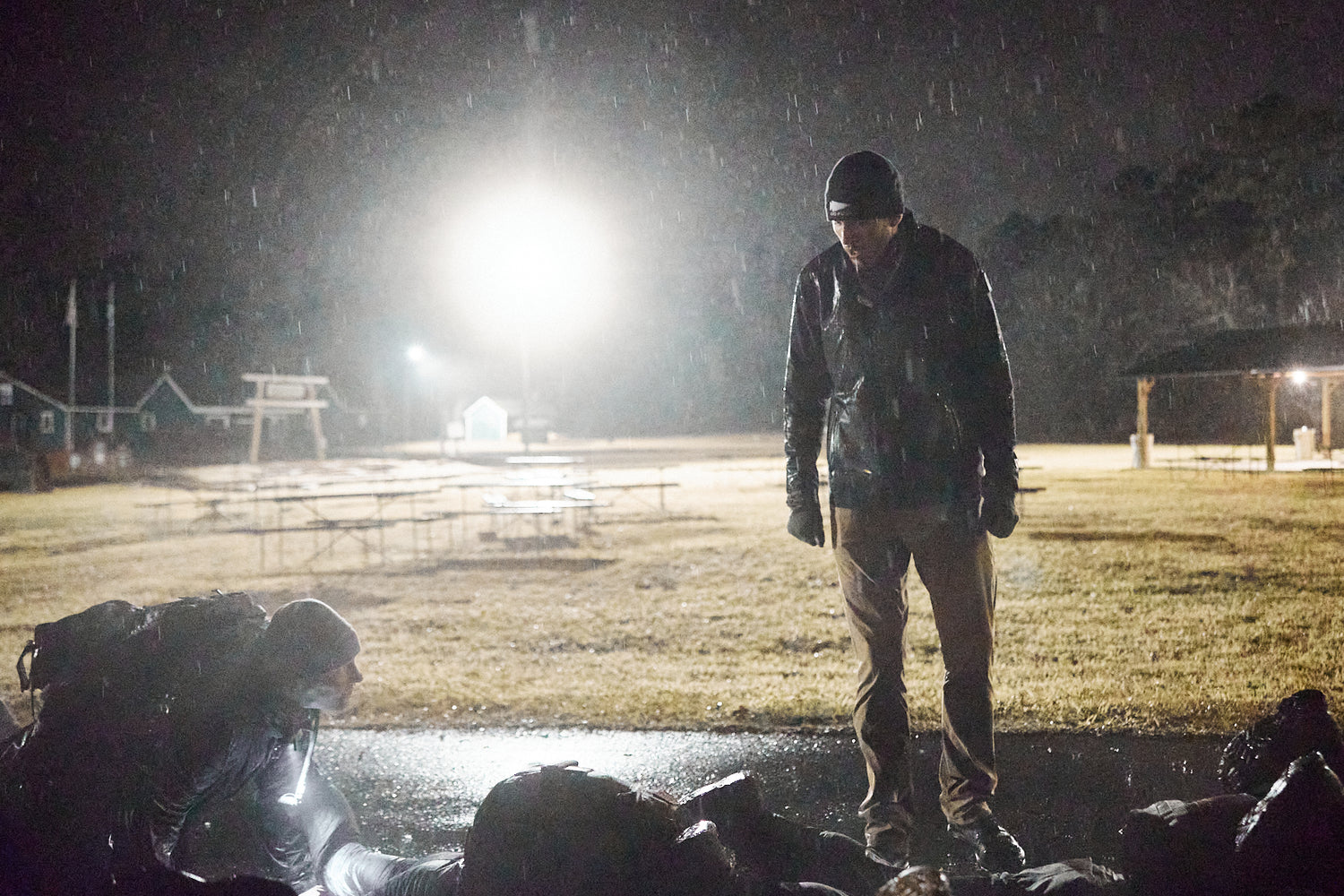 A person stands over others lying on wet ground at night, with a bright light shining in the background.