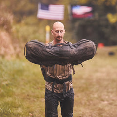 A man lifts a heavy sandbag outdoors using the Padded Hip Belt for support, with blurred American flags in the background.