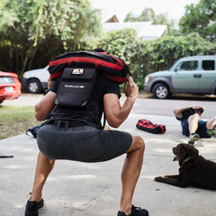 A person does squats outdoors with a weighted backpack in Men’s Simple Shorts - Lightweight ToughDry®, while a dog and another person rest nearby.