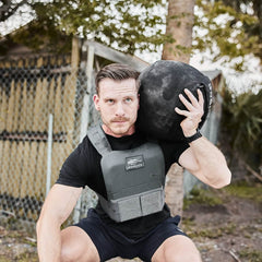 A man squats outdoors wearing the Training Weight Vest 2.0, holding a large black medicine ball on his shoulder.