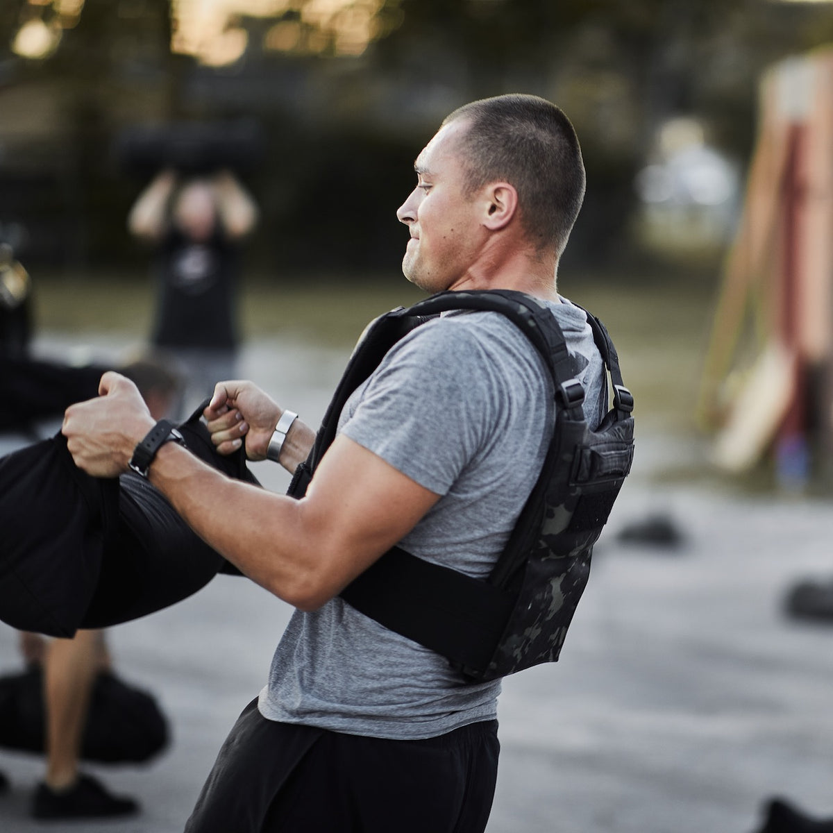A man wearing a training weight vest lifts a black sandbag during an outdoor fitness workout.