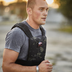 A man in a grey t-shirt runs wearing a black camouflage GORUCK training weight vest with an American flag patch.
