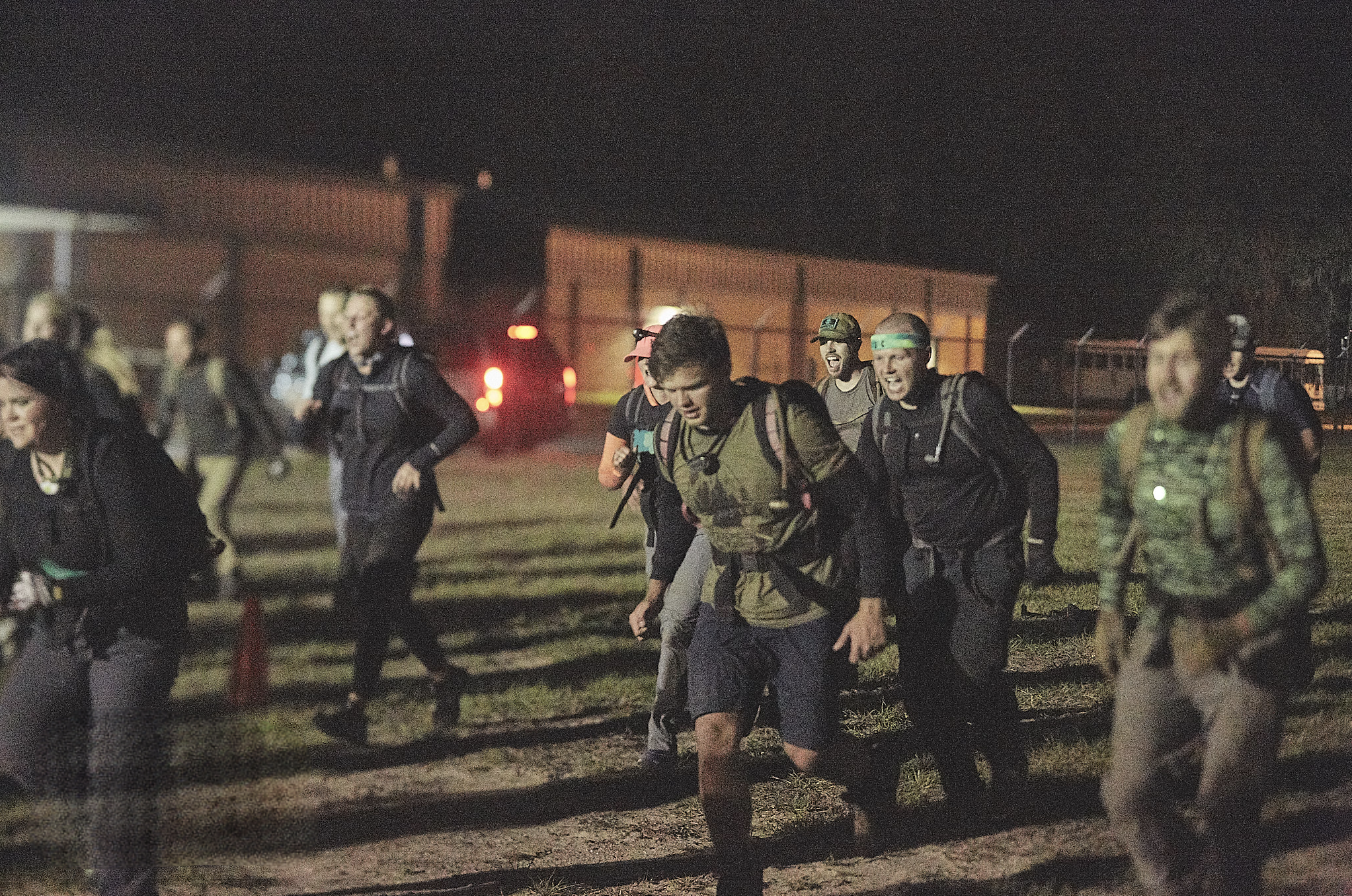 People wearing backpacks and athletic gear running outdoors at night, illuminated by artificial lights.