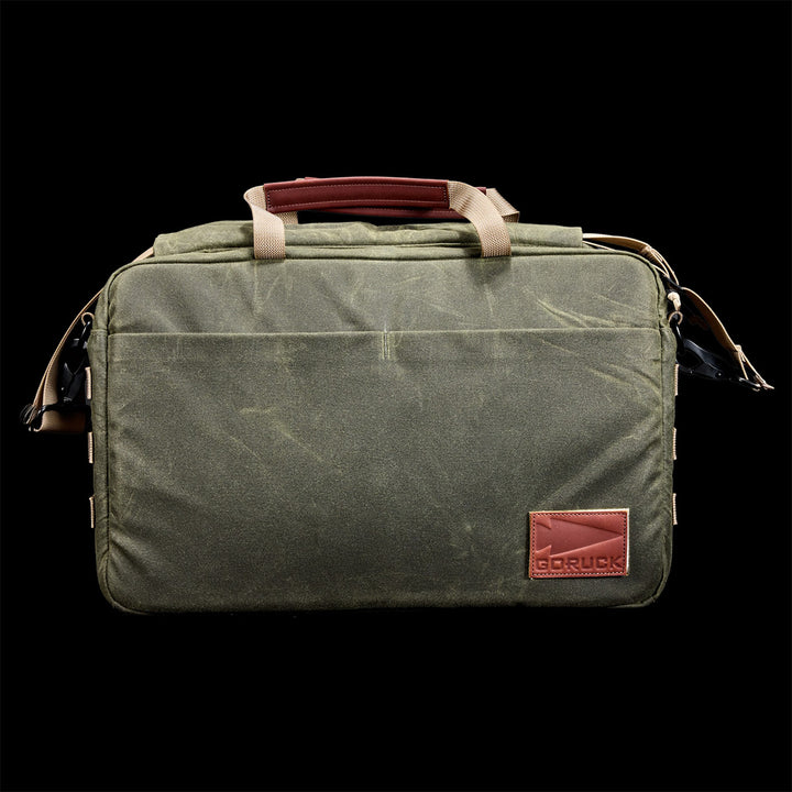 The Shoulder Bag - Waxed Canvas features olive green canvas, tan handles, a brown leather GORUCK logo patch, and a removable strap for versatile carry, shown against a black background.