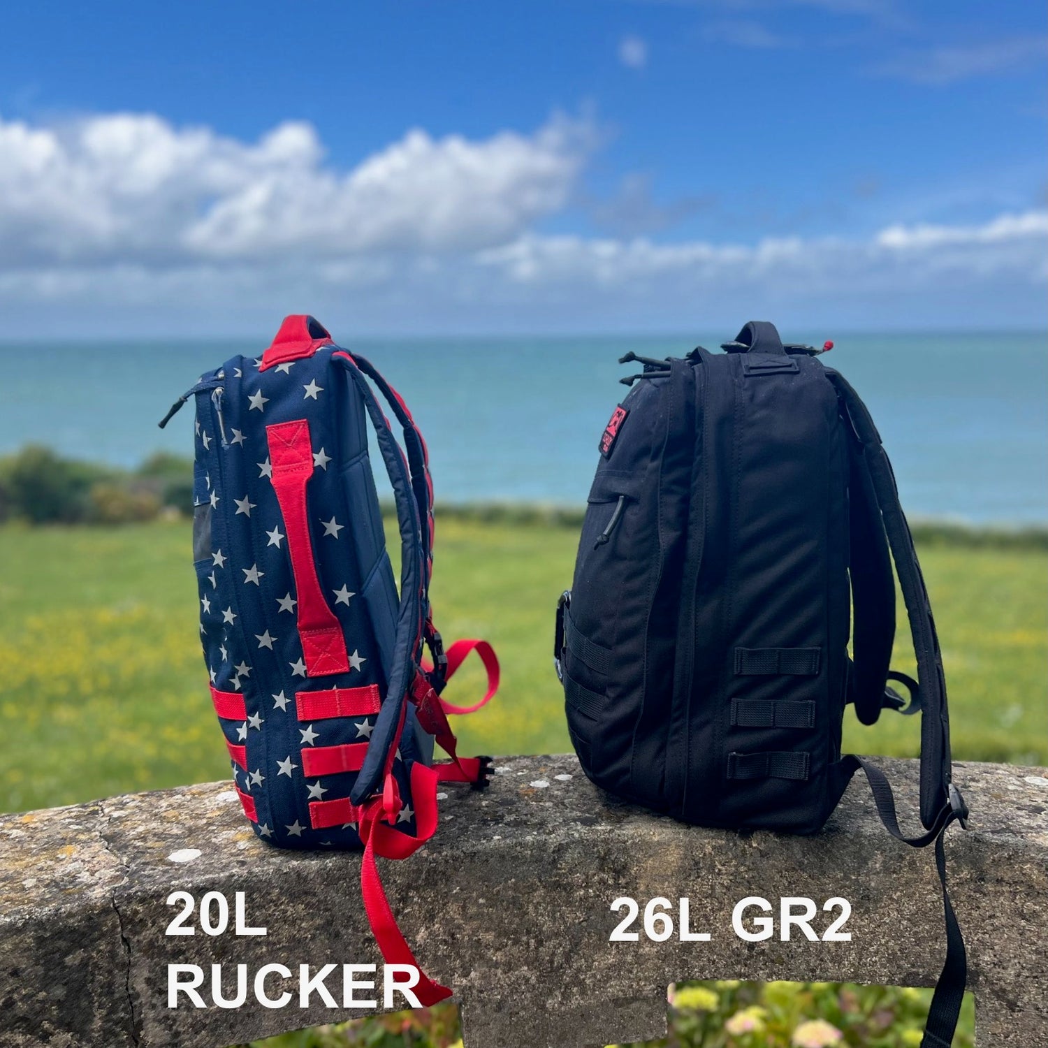A black 26L GR2 ruck and a star-patterned 20L Rucker sit on a stone wall by the sea, perfect for one bag travel adventures.