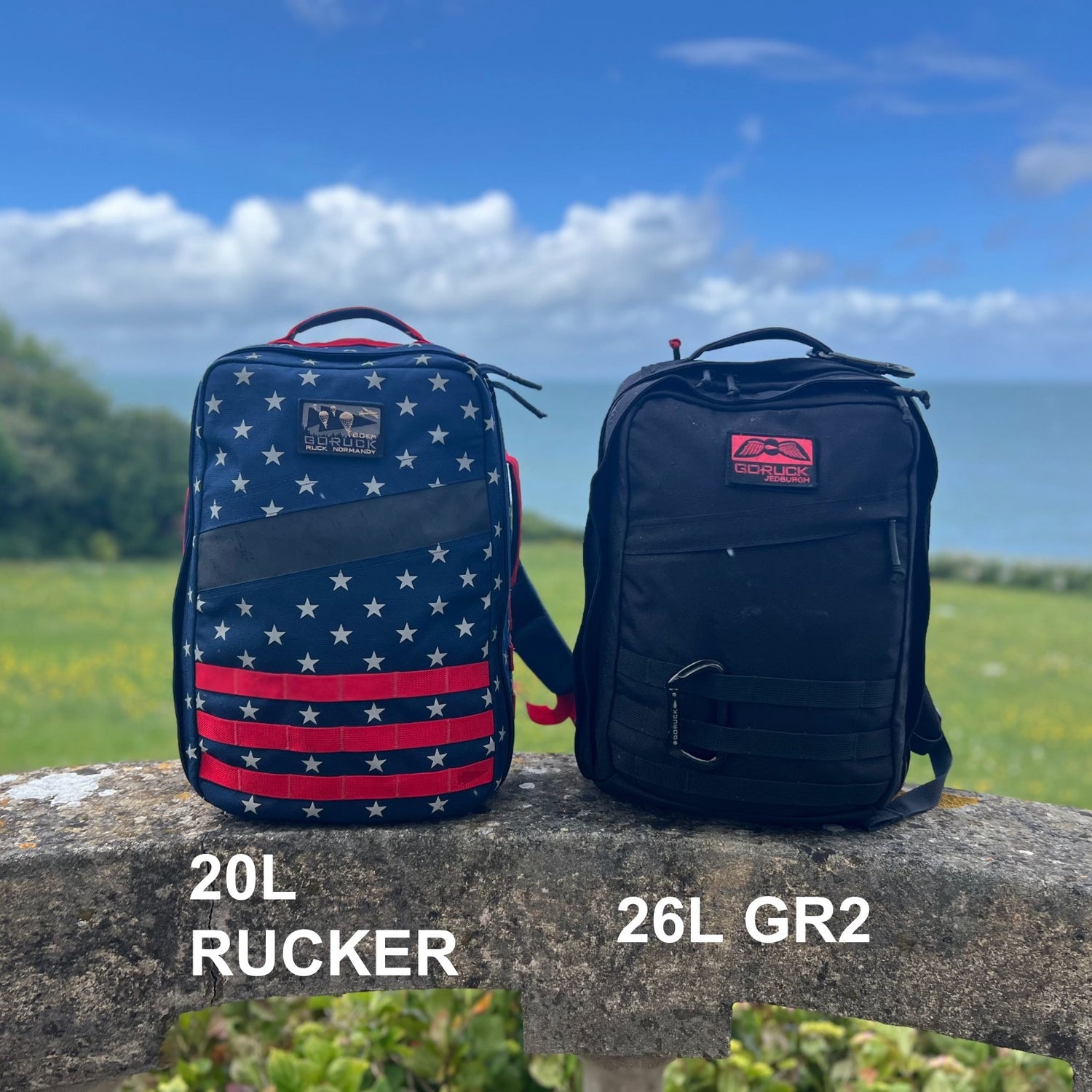 Two GR2 backpacks—one with an American flag pattern and one in black—sit on a stone ledge by the ocean, ideal for one bag travel adventures.