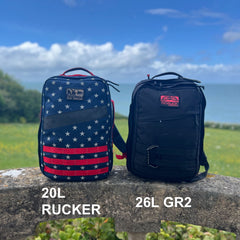 Two GR2 backpacks—one with an American flag pattern and one in black—sit on a stone ledge by the ocean, ideal for one bag travel adventures.