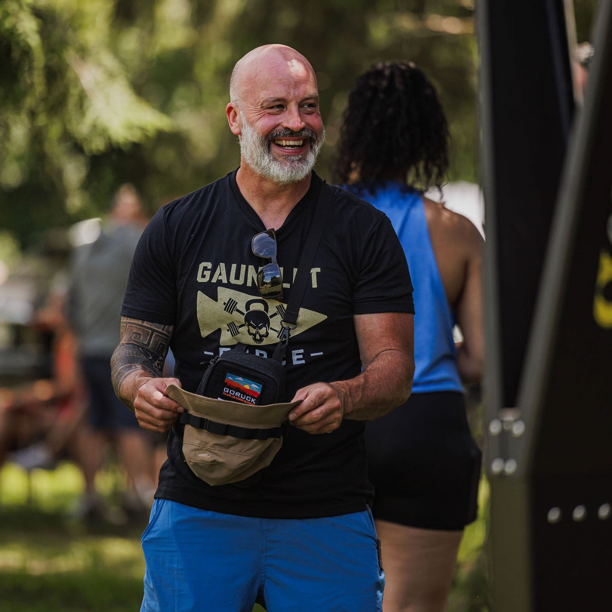 Bald man with a beard smiles outdoors, holding the Boonie Hat - Tactical - ToughDry, dressed in a black t-shirt and blue shorts for a relaxed military look.