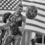 A man covered in mud throws a Sand Medicine Ball with a large American flag in the background, demonstrating core stability and strength.