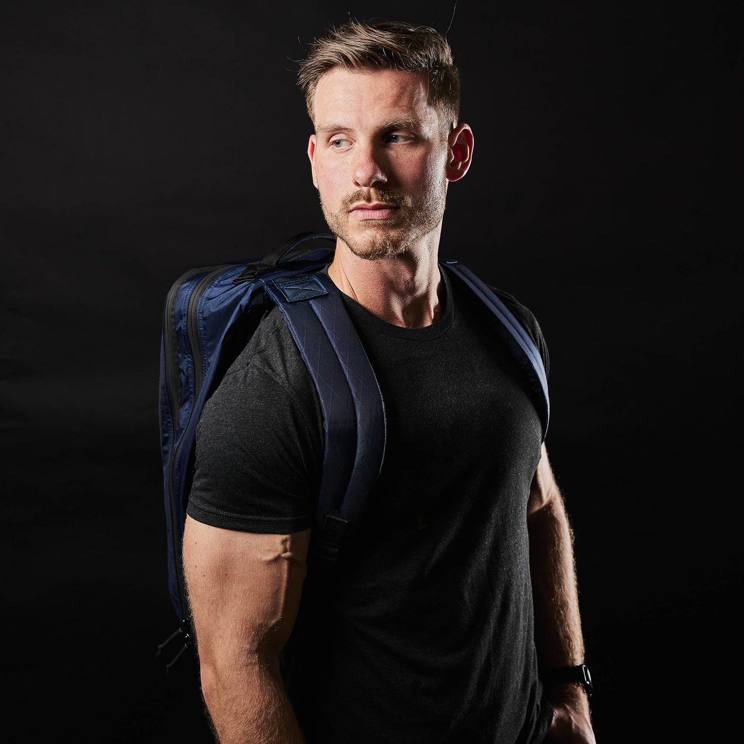 A man with short hair and a beard wearing the Slick Short Sleeve Tee - Tri-BlendX in black, featuring antimicrobial and premium tri-blend fabric, paired with a blue backpack and looking to the side against a black background.