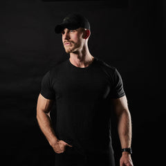 A man wearing the Slick Short Sleeve Tee - Tri-BlendX in black and a cap stands against a dark background, looking slightly to the side.