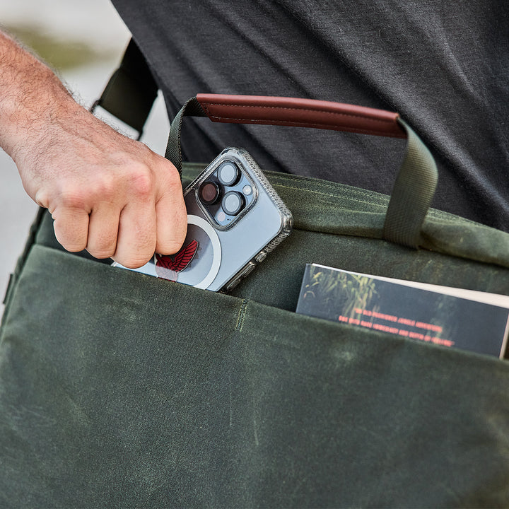 A person carefully slips a smartphone into the pocket of a green waxed canvas bag, nestled alongside a book with its cover partially visible. This chic Shoulder Bag - Waxed Canvas by GORUCK seamlessly merges functionality and style, ideal for those who love asymmetrical carrying options while on the move.