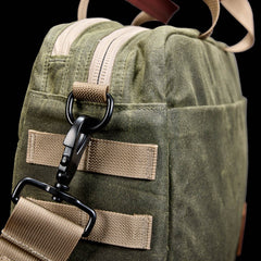Close-up of the GORUCK Shoulder Bag - Waxed Canvas in olive green with beige straps, a black metal clip, and Martexin Wax finish, designed for comfortable asymmetrical carries against a black background.