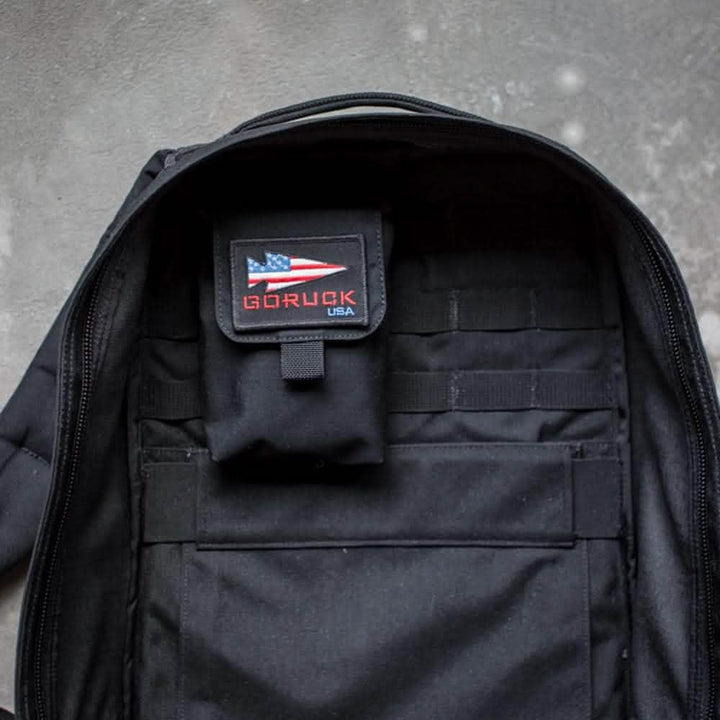 An open black rucksack reveals a sewn-in Simple Side Pocket featuring a sleek pouch with the GORUCK USA label and an American flag patch. The design incorporates MOLLE webbing for versatile use, and the backpack rests on a gray surface.