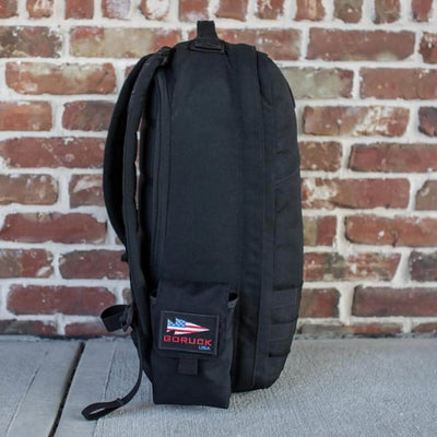 A black backpack with MOLLE webbing and the Simple Side Pocket, adorned with a USA flag patch and GORUCK branding, stands upright on a concrete surface against a red brick wall.