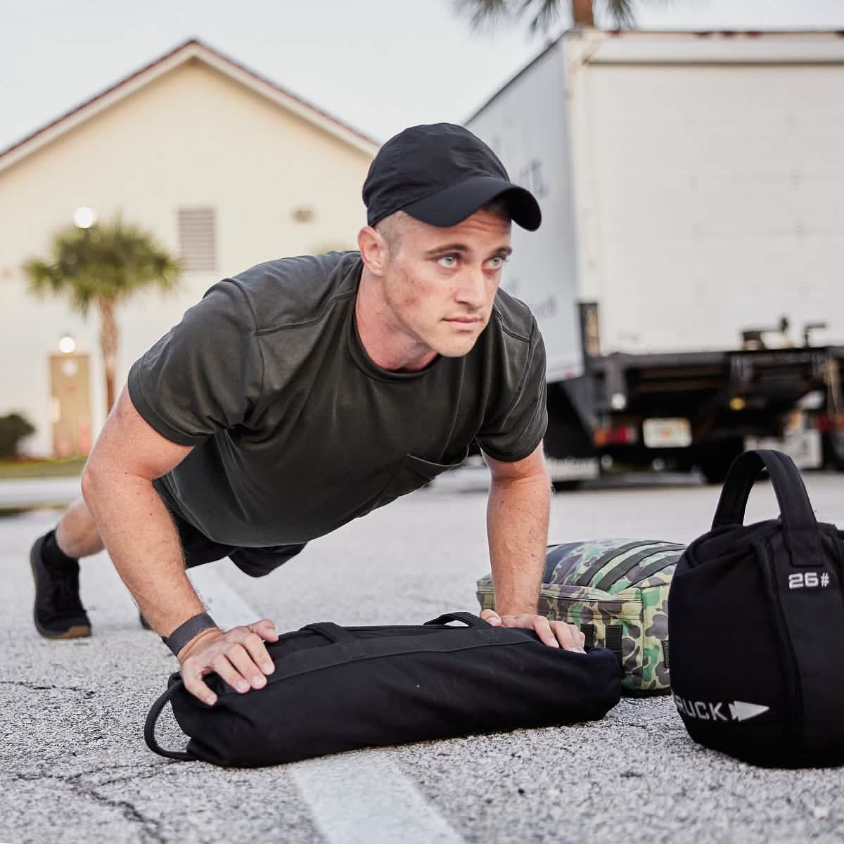 A man wearing a black Performance TAC Hat - Slick by GORUCK and a t-shirt is doing push-ups on a street, beside weighted bags and a rucksack. The TOUGHDRY fabric of his attire helps him stay dry while he trains. A truck and a house are in the background, completing the rugged scene.