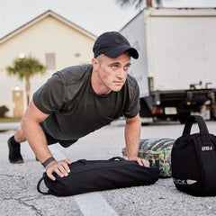 A man wearing a black Performance TAC Hat - Slick by GORUCK and a t-shirt is doing push-ups on a street, beside weighted bags and a rucksack. The TOUGHDRY fabric of his attire helps him stay dry while he trains. A truck and a house are in the background, completing the rugged scene.