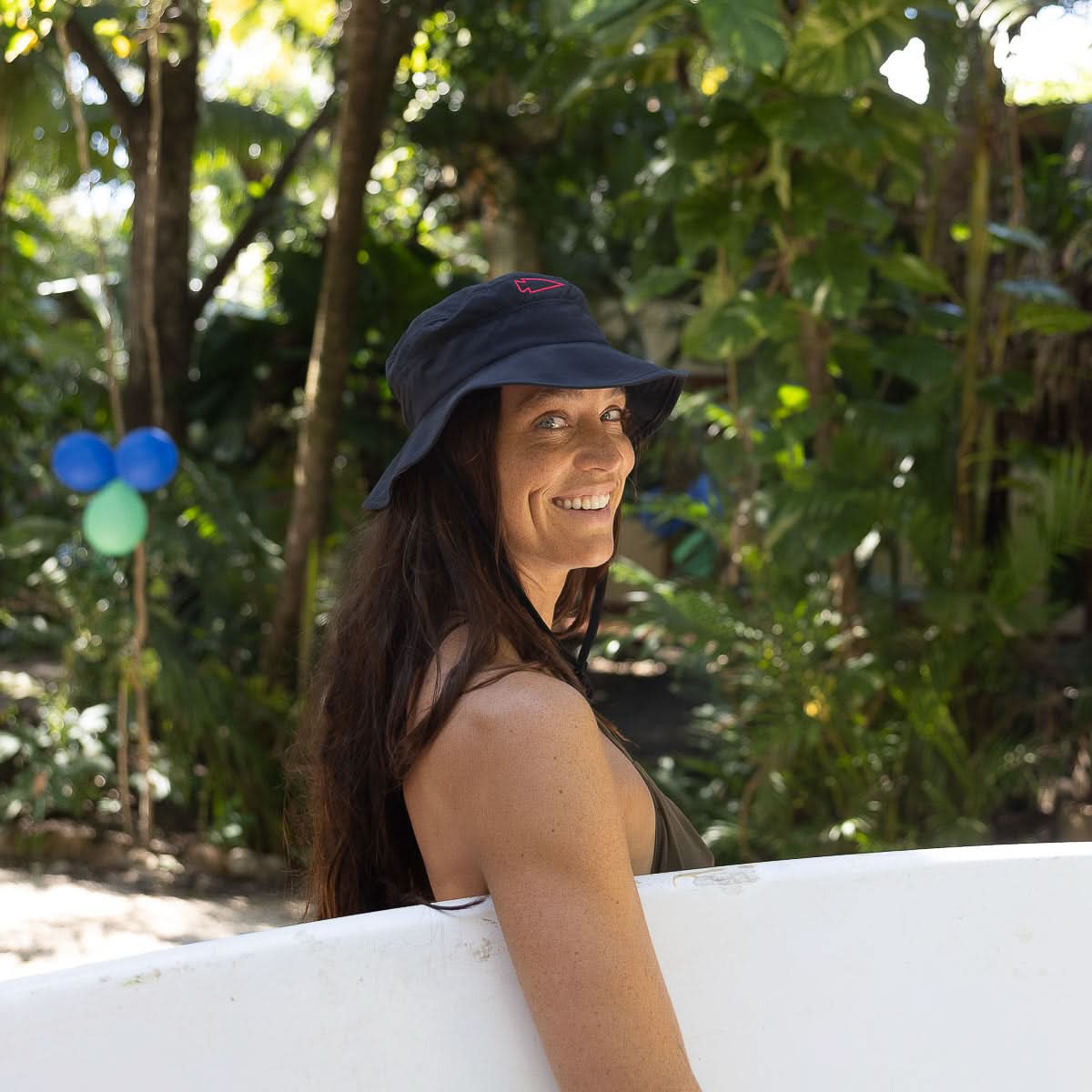Woman wearing a Boonie Hat - Slick - ToughDry holding a surfboard, smiling among tropical balloons and greenery.
