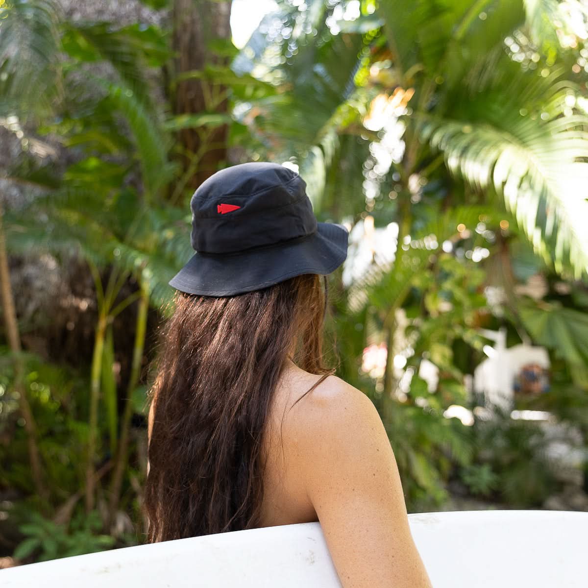 A person with long hair, wearing a black Boonie Hat - Slick - ToughDry, stands before lush greenery holding a surfboard.