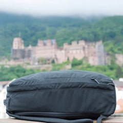 A GR1 USA Slick - Ballistic Nylon Cordura backpack rests on a ledge, its bombproof laptop compartment ready for adventure, with a blurred castle and green hills in the background.