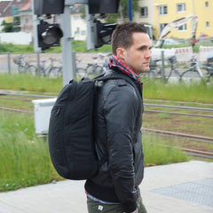 A man with a black GR1 USA Slick - Ballistic Nylon Cordura backpack waits by tram tracks, wearing a black jacket and plaid shirt—his bag’s rugged build ensures bombproof laptop protection for every adventure.