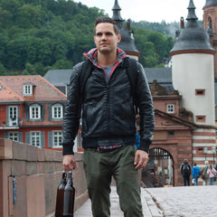 A man in a black jacket walks on a bridge, carrying bottles and the GR1 USA Slick - Ballistic Nylon Cordura backpack; historic buildings and trees create a scenic backdrop.