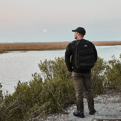A person stands on a shoreline, donning the GORUCK Performance TAC Hat in Slick and a jacket, gazing at the water and marshland. The sky is overcast, with hints of the setting sun on the horizon. Bushes frame this tranquil scene as TOUGHDRY® fabric provides protection against the elements.