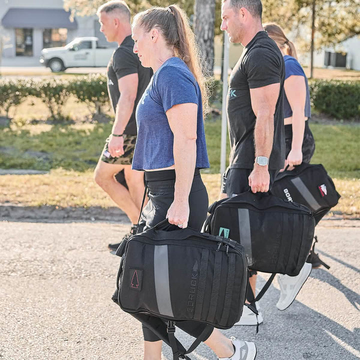 Four individuals walking outdoors, dressed in athletic gear, each carrying a large backpack decorated with an embroidered patch featuring the Spearhead Outline from GORUCK. They stroll along a paved path with grass and trees nearby, under a clear sky.