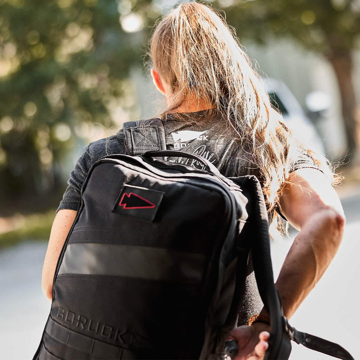 A person with long hair carries a black backpack adorned with GORUCK's Spearhead Outline patch while outdoors. The individual is wearing a gray shirt and viewed from behind, as sunlight filters through the trees, casting soft shadows on the ground.