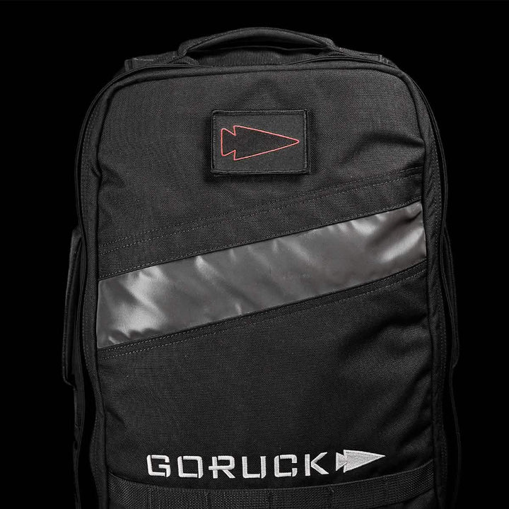 A black GORUCK backpack adorned with the Patch - Spearhead Outline, which includes an embroidered patch featuring a diagonal reflective stripe and VELCRO® backing, highlighted by a red arrowhead design. The iconic GORUCK logo is prominently displayed at the bottom in crisp white text.