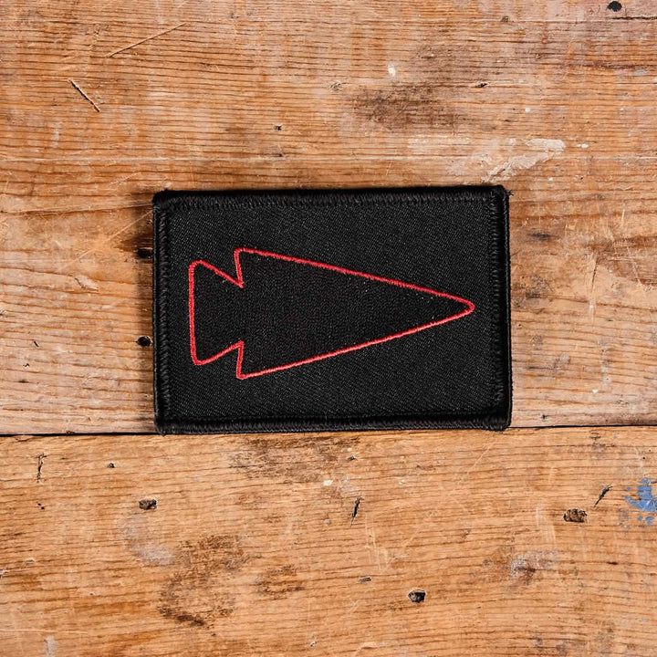 A black embroidered GORUCK patch, featuring the Spearhead Outline design in red, rests on a wooden surface. The rustic wood, with its textured appearance and visible grain, complements the patch's intricate details.