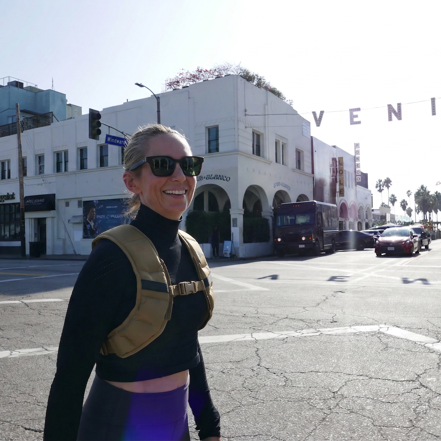A smiling woman in sunglasses and a Spy Ruck Women's Weighted Vest crosses a sunny street near the Venice sign in an urban area.