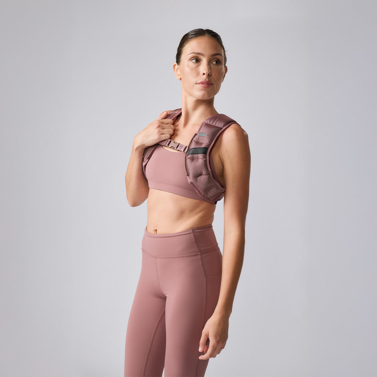 A woman in pink athletic wear and a Spy Ruck weighted vest stands confidently against a plain light gray background, highlighting top women’s fitness gear.