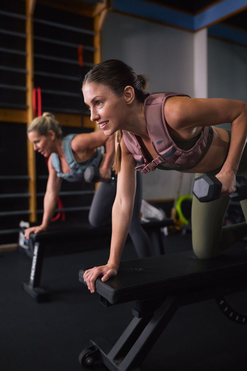 Two women lifting dumbbells while leaning on workout benches in a gym, focused on their exercise.