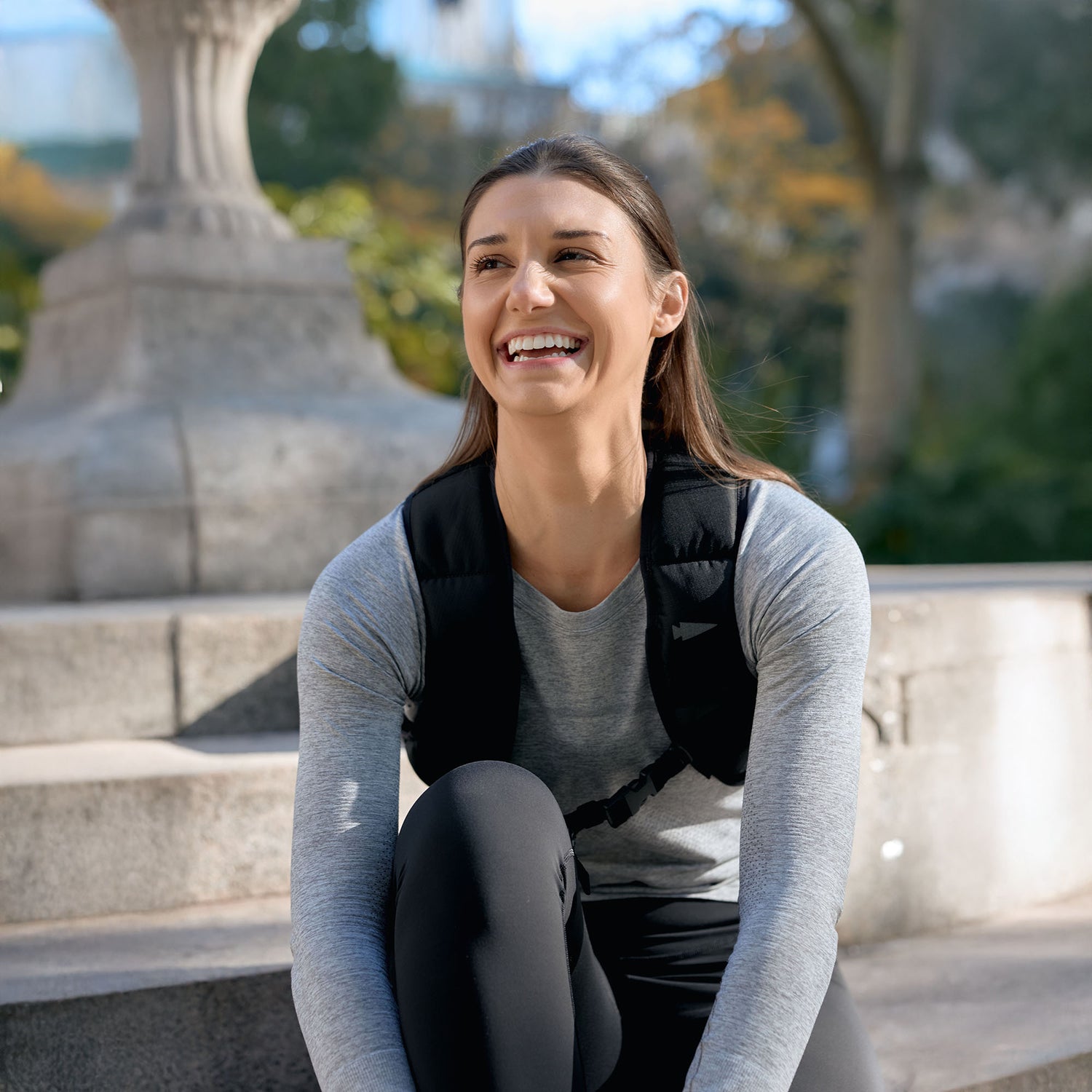 A smiling woman in athletic wear and the Spy Ruck | Women's Weighted Vest sits on outdoor stone steps with trees in the background.