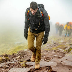 A hiker wears T-REX Hand Straps (pair) to reduce hand swelling while climbing a rocky, foggy trail, with a group following behind.