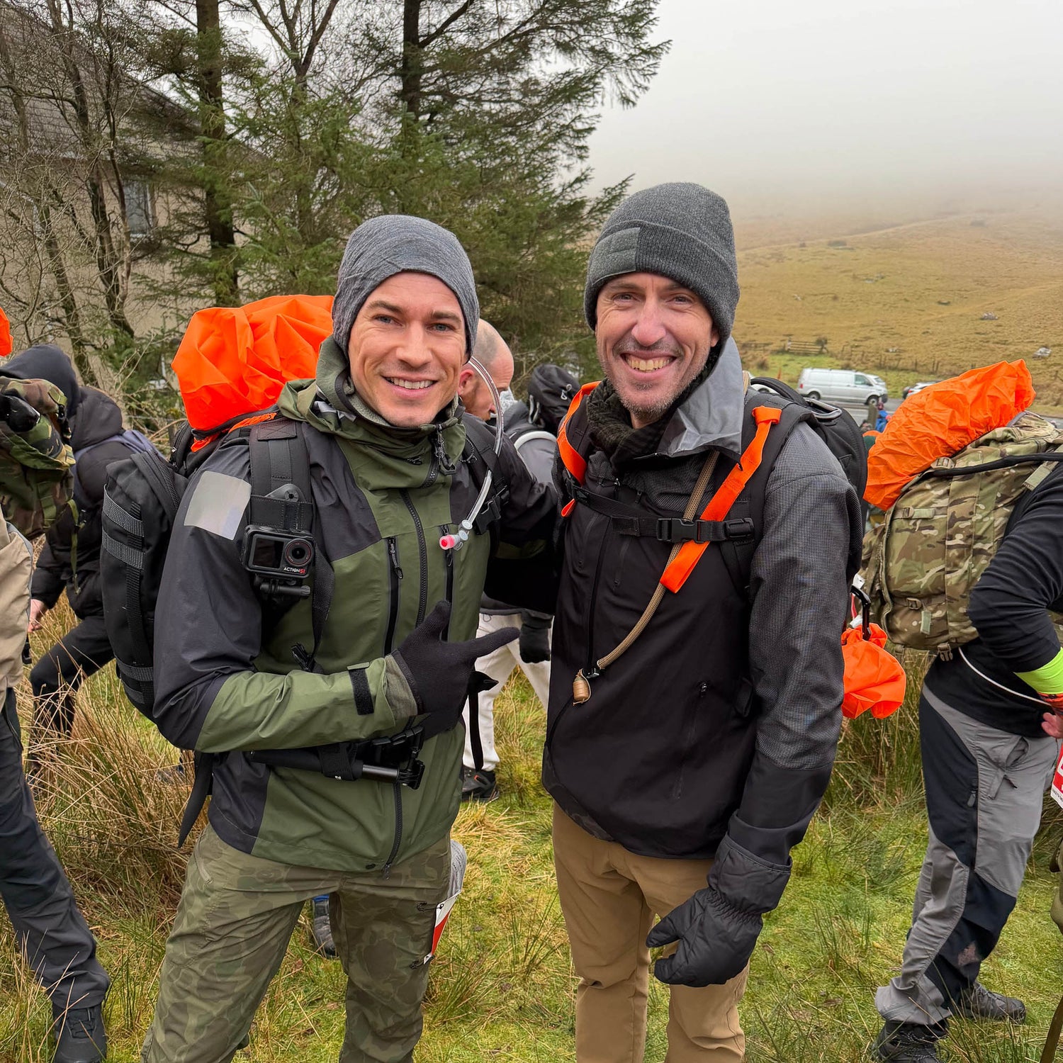 Two men in outdoor gear and backpacks, wearing T-REX Hand Straps (pair) for hand swelling relief, smile on a foggy, grassy hillside with others visible in the background.