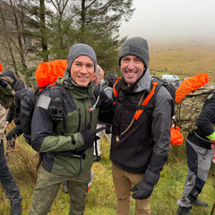 Two men in outdoor gear and backpacks, wearing T-REX Hand Straps (pair) for hand swelling relief, smile on a foggy, grassy hillside with others visible in the background.