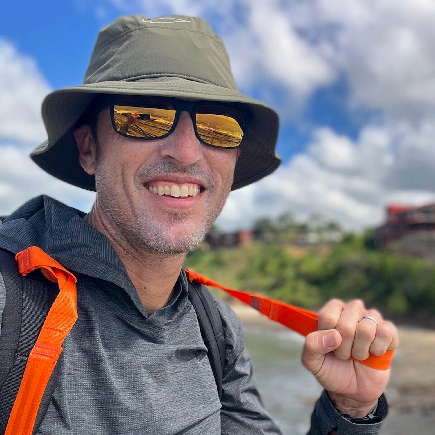 A man in a hat and sunglasses, smiling outdoors with a backpack and T-REX Hand Straps (pair)—ideal rucking gear for hand swelling relief—stands against a blurred nature background.