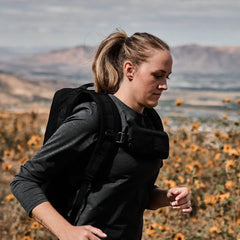 Woman rucking outdoors wearing a black sternum pouch and backpack, running in a field with mountains in the background