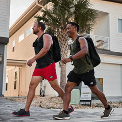 Two men carrying black rucksacks walking outdoors near modern homes with palm tree, wearing athletic gear