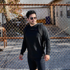 Dressed in a sleek GORUCK Subtle Spearhead Hoodie - Tri-Blend and sunglasses, the man stands confidently by the chain-link fence, his tri-blend backpack casually slung over one shoulder.