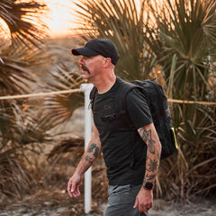 A tattooed man with a mustache walks outdoors at sunset, wearing the Subtle Spearhead Tee - Tri-Blend, surrounded by palm plants.
