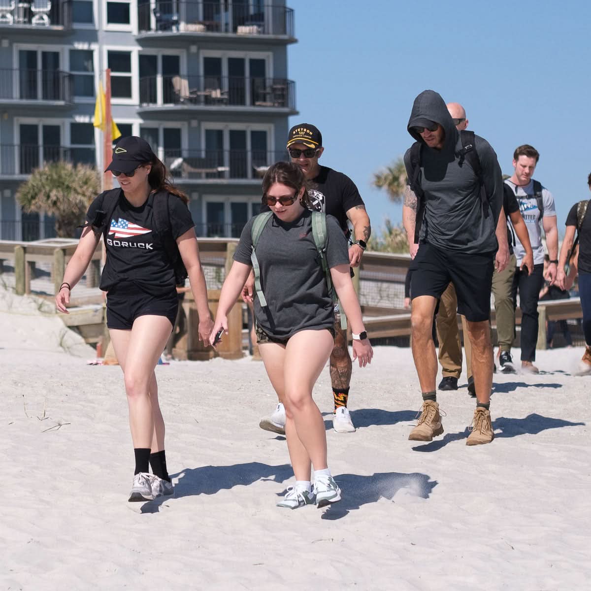 A group wearing Subtle Spearhead Tee - Tri-Blend shirts, some showing off the GORUCK spearhead logo, walk together on a sandy beach near a building.