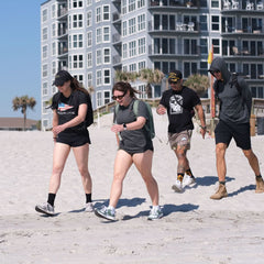 Four people in tri-blend athletic wear stroll on a sandy beach near modern apartments, with one wearing the Subtle Spearhead Tee - Tri-Blend from GORUCK.